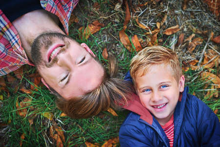 He is his fathers son. High angle portrait of a handsome young man and his son lying on the grass outside during autumn.の写真素材