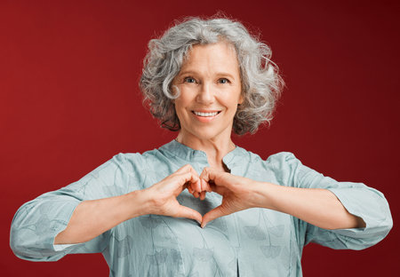Love, heart and health retired woman with loving emoji sign, icon or symbol showing care or affection on red valentines day studio background. Grey, senior or pensioner with a trendy emoticon poseの写真素材
