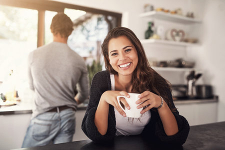 Coffee is cuppa happiness. Portrait of a young woman having coffee in the kitchen with her husband standing in the background.の写真素材