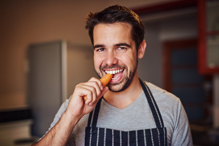 Carrots are perfect for a crunch craving. Portrait of a happy young man eating a carrot while standing in his kitchen at home.の写真素材
