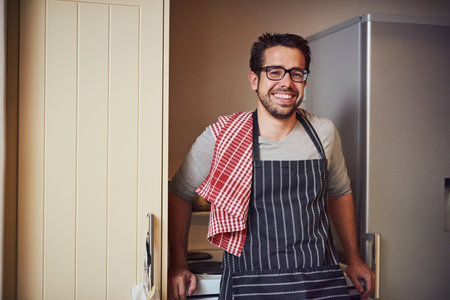 Good food, great mood. Portrait of a happy young man wearing an apron while posing in his kitchen at home.の写真素材