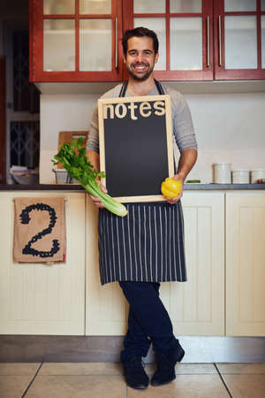 The kitchen is where adventures happen. Portrait of a happy young man posing with fresh ingredients and a blank chalkboard in his kitchen.の写真素材