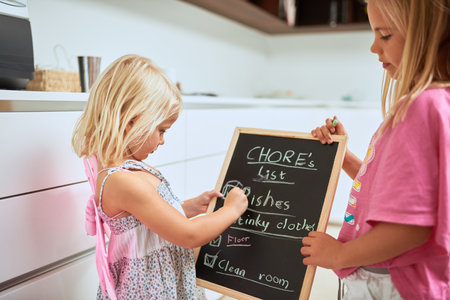 Theyre always willing to help with housework. two little girls writing a list of chores on a chalkboard at home.の写真素材