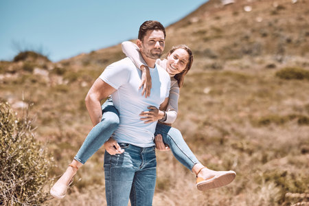 Happy and in love couple having fun in nature and enjoying quality time together. Cute and relax boyfriend piggyback or carrying girlfriend with a smile on a bonding and romantic countryside dateの写真素材