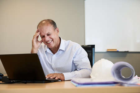 He is happy and excited about the things to come. Portrait of a cheerful professional businessman working on his laptop while having a laugh inside the office.の写真素材