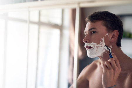 Ill face the day with a smooth face. a young man shaving his beard while looking at himself in the mirror.の写真素材