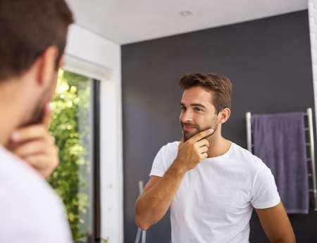 To shave or not to shave thats the question. a handsome man rubbing his beard and looking in the mirror at home.の写真素材