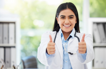 Excited medical doctor showing thumbs up for success working at hospital, giving support after and happy with service in an office at clinic. Portrait of healthcare worker showing winning hand signの写真素材