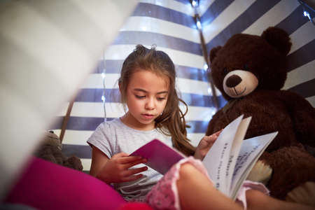 Shes a keen reader. a little girl reading a book with her teddybear in a tent at home.の写真素材