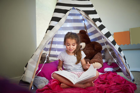 Books can help fuel a childs curiosity and imagination. a little girl reading a book with her teddybear in a tent at home.の写真素材