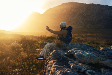 The mountains are calling me, I must answer. a hiker sitting on top of a mountain capturing photos with his cameraphone.の写真素材