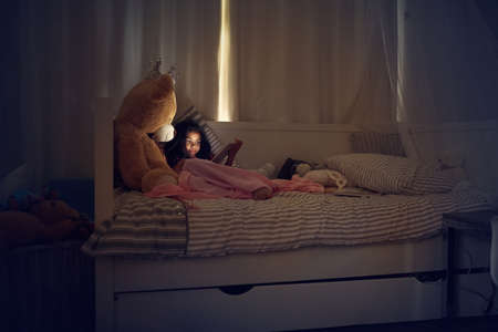 Technology keeps her entertained day and night. a little girl using a digital tablet while lying in bed with her teddy at night.の写真素材
