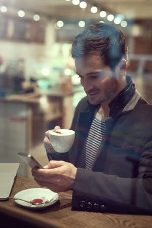 Im grabbing a coffee quick. a handsome young man using his cellphone while sitting in a cafe.の写真素材