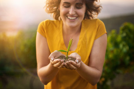 Growing up green. a happy young woman holding a small seedling in her cupped hands while standing outside.の写真素材