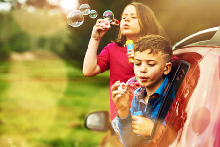 The more bubbles, the better. two happy children blowing bubbles while leaning out of a car window together.の写真素材