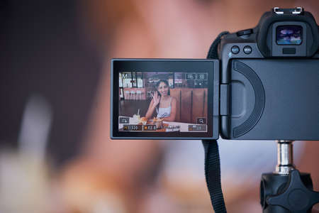 Woman influencer on digital camera screen recording vlog for online social media video. Creative young female content creator presenting internet or web content for her website or podcast at a cafeの写真素材