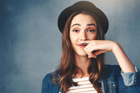 Lets all support Movember. Studio shot of an attractive young woman making a finger moustache against a blue background.の写真素材