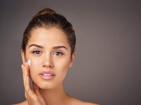 Nourished skin is beautiful skin. Studio portrait of a beautiful young woman applying moisturizer to her face against a gray background.の写真素材