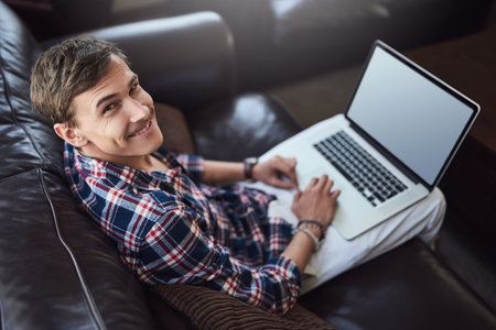 This blogging thing is kinda fun. High angle portrait of a handsome young man using his laptop while sitting on the sofa at home.の写真素材