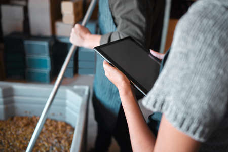 Owner working on a digital tablet in a winery while a wine maker press grapes to make wine. Woman...