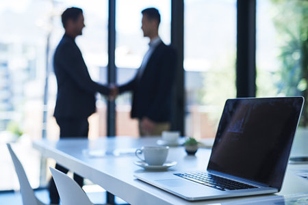 Taking business to the next level. a laptop on a table in an office with two businessmen shaking hands in the background.の写真素材