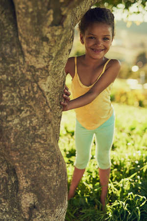 No one will find me here. Portrait of a smiling girl playing outside behind a tree.の写真素材