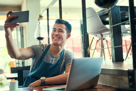 Selfies at work. a young man taking selfies while working in his coffee shop.の写真素材