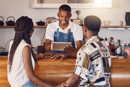 Hospitality, service and technology with restaurant worker taking orders on a digital tablet, smiling and talking. Young employee manage customer payment on an online app, paying using the internetの写真素材