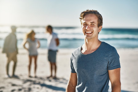 The beach is our happy place. Portrait of a happy young man posing on the beach with his friends in the background.の写真素材