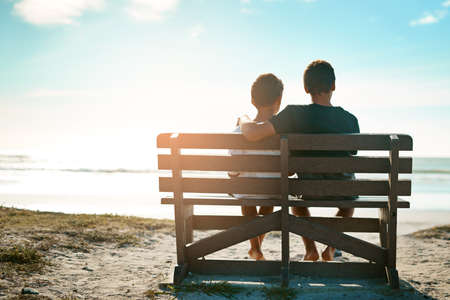 Well be best friends for life. Rearview shot of two unidentifiable brothers sitting on a bench together by the beach.の写真素材