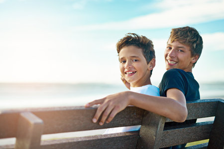 Theres no sibling rivalry here. Portrait of two happy brothers sitting on a bench by the beach together.の写真素材