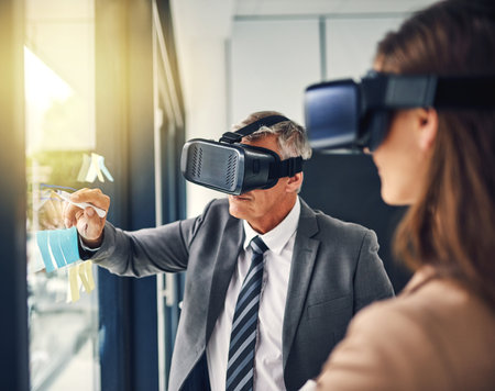 Reaching out to where it was thought impossible before. two businesspeople wearing VR headsets while working with notes on a glass wall in an office.の写真素材