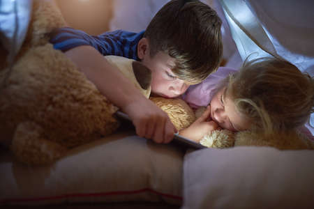Technology is there to entertain them day and night. siblings using a digital tablet under a blanket fort at home.の写真素材