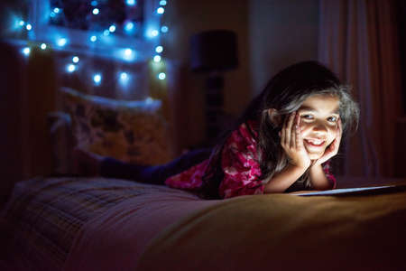 My bedtime stories glow in the dark. Portrait of a little girl using a digital tablet late at night in her bedroom.の写真素材