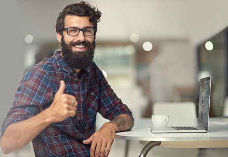 I enjoy the feeling of accomplishment after a project. a young designer showing thumbs-up while sitting by his laptop.の写真素材