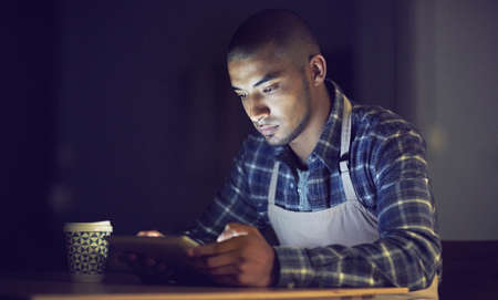 Putting in the hours. a young man working late on a digital tablet in his coffee shop.