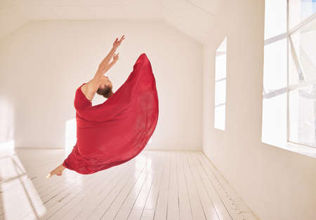 Woman, ballet and dance of a girl student dancing in a red dress in a studio mockup with white walls and sunlight. Young professional female ballerina, art or dancer jumping in the air in a class.の写真素材