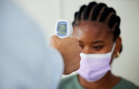 Covid, healthcare and woman patient getting thermometer check with a compliance officer during consultation or appointment. Health, hygiene regulations and wellness with a female wearing a face maskの写真素材