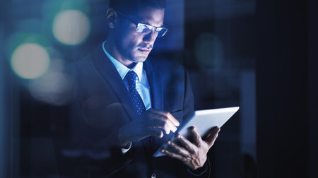 Businessman working overtime on a digital tablet with glasses in a corporate modern office. Corporation manager reading company documents, data and statistics on an internet website with technology.の写真素材
