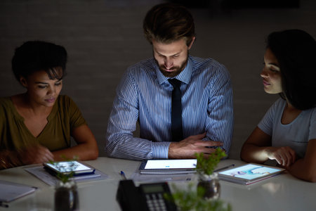 Working late to get the job done. a group colleagues using digital tablets while working late in an...