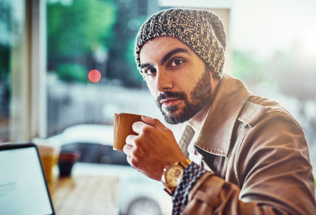 He takes his coffee seriously. Portrait of a handsome young man sitting at a cafe counter using a laptop and drinking a cup of coffee.の写真素材