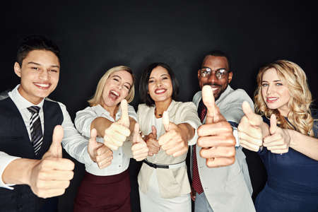 We all think youre doing a great job. Portrait of a group of businesspeople showing thumbs up against a dark background.の写真素材