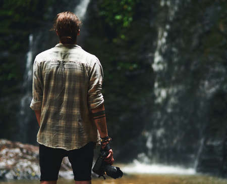 This is amazing. a young man staring at a waterfall with his back turned and holding his camera.の写真素材