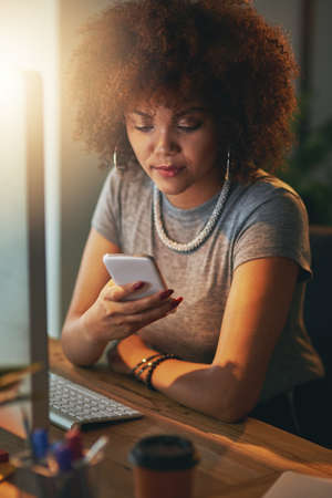 Getting the job done with hard work and extra hours. a young woman using a phone during a late shift at the office.の写真素材