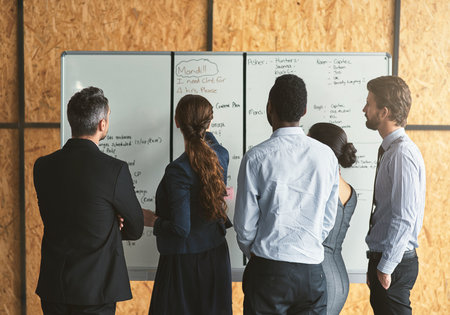 Planning the way forward. Rearview shot of a group of businesspeople looking over plans on a whiteboard.の写真素材