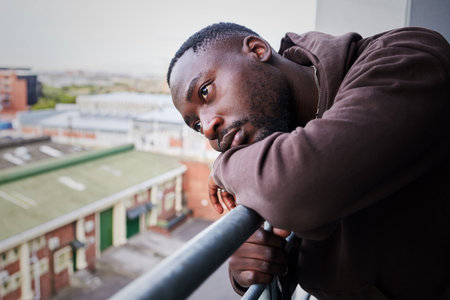 Sad, tired and depressed man on balcony of apartment, thinking of mental health in house and anxiety from problem at home. Face of African person with depression and burnout thinking of ideaの写真素材