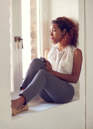 Shes got a busy day ahead of her. a young woman using her cellphone while sitting by a window.の写真素材