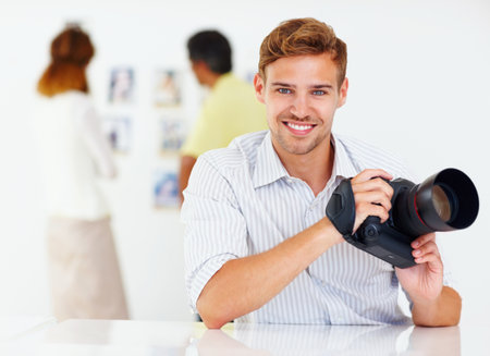 Young photographer with camera. Portrait of young photographer holding camera and smiling with colleagues in background.の写真素材