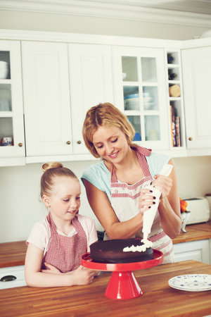 Piping on the final finishings. Cute little girl icing a cake with her mom in the kitchen.の写真素材