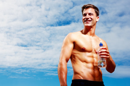 Happy young man looking away with water bottle. Portrait of a happy young man looking away while holding a bottle of water against sky.の写真素材